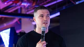 A man on a stage in a black T-shirt speaking into a microphone.