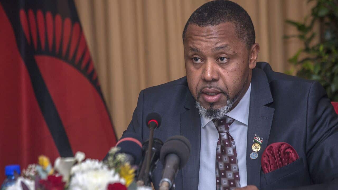 Malawian vice president Saulos Klaus Chilima sits behind a cluster of microphones and a bouquet of flowers. He is wearing a dark blue suit.