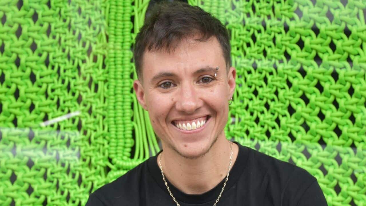 A person with short brown hair and wearing a black T-shirt smiles for a photo in front of a dangling sheet of bright green crocheted fabric.