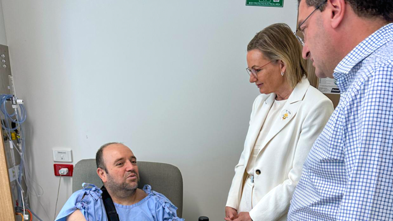 Ahmed Al-Ahmed in a hospital chair, smiling as Sussan Ley and Julian Leeser look on