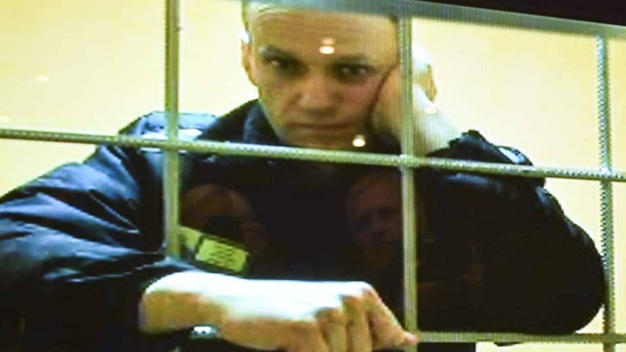 A man with short hair wearing a prison jumpsuit looks through the bars of a prison cell.