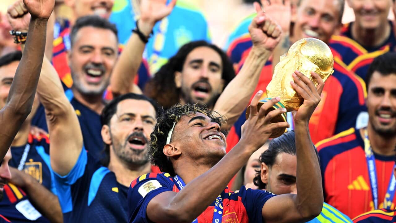 A smiling young soccer player with curly hair and braces joyfully hoists the golden FIFA World Cup trophy as his teammates celebrate around him.