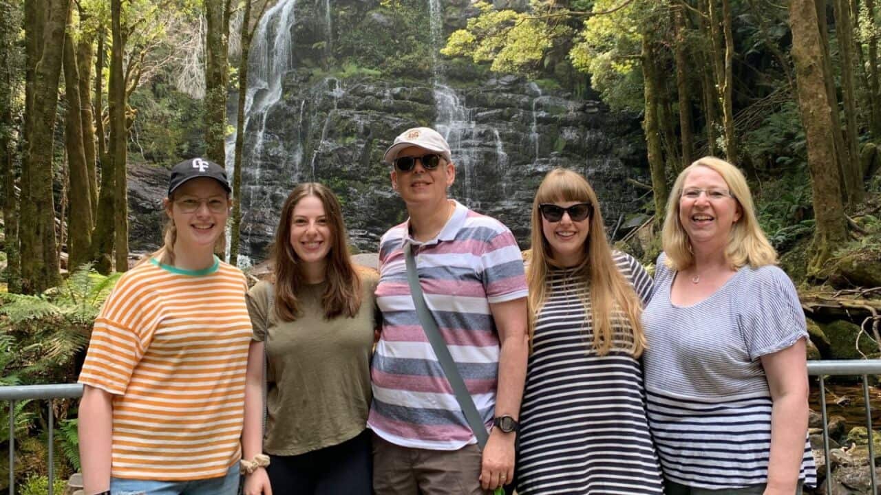 a family photo in front of a waterfall - one young women, a middle aged man, another young woman and a middle aged woman stand close together smiling