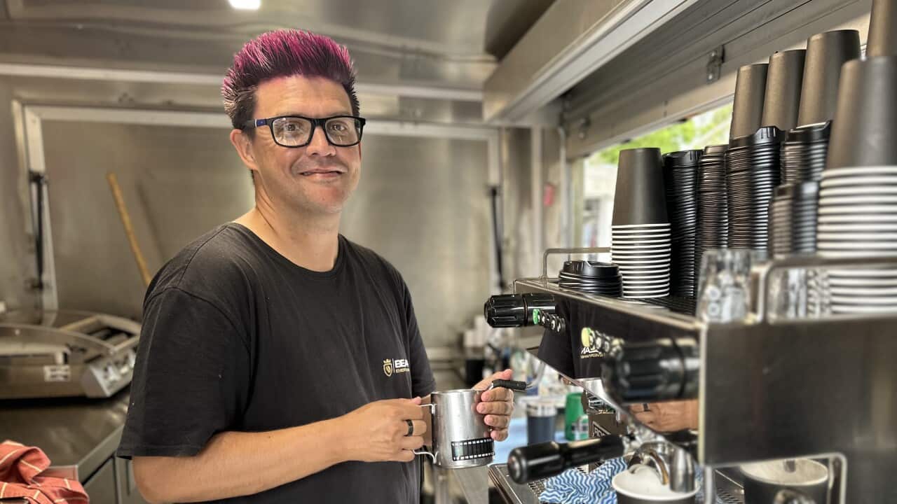 A barista in a silver coffee van smiles to camera while making a coffee
