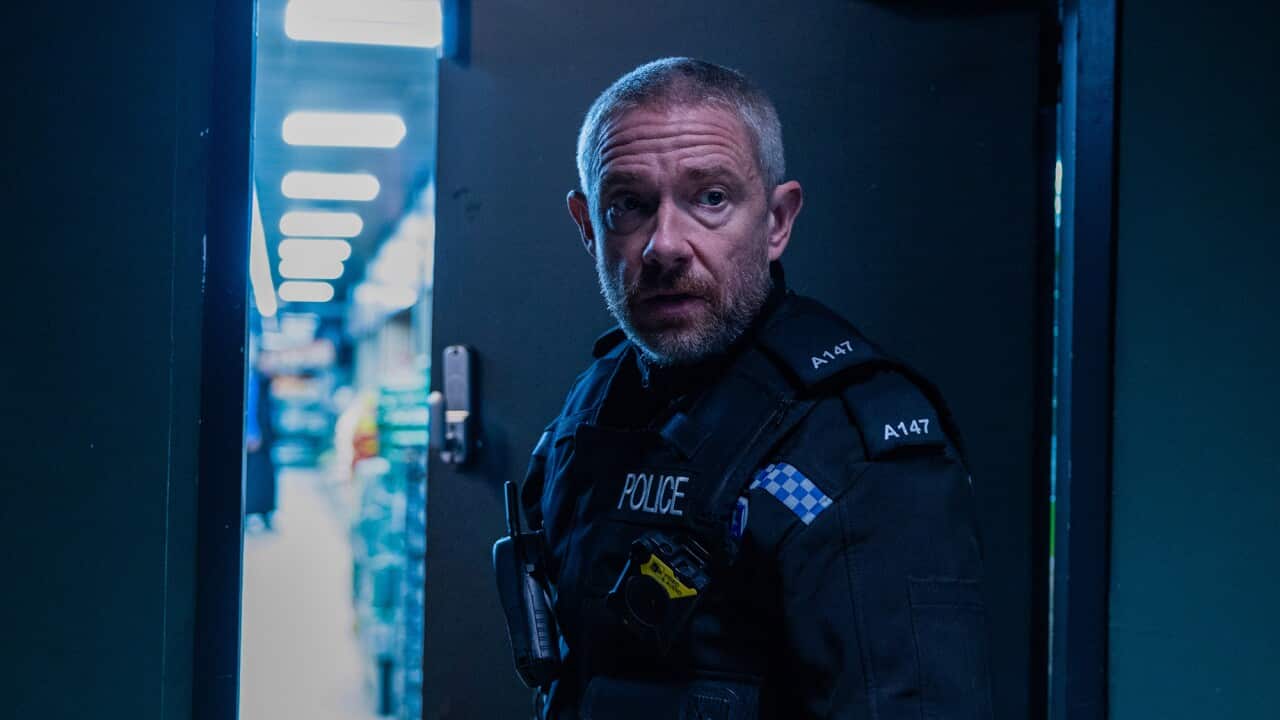 A man in a police uniform stands in a dark doorway, with a well-light rooom partly seen through behind him.