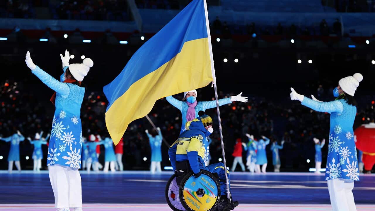 A wide shot of the Ukrainian delegation during the opening ceremony of the 2022 Winter Paralympics. At the center, an athlete in a wheelchair wears a blue and yellow team uniform. He holds a tall flagpole, and the large Ukrainian flag (blue and yellow) waves prominently above him.
