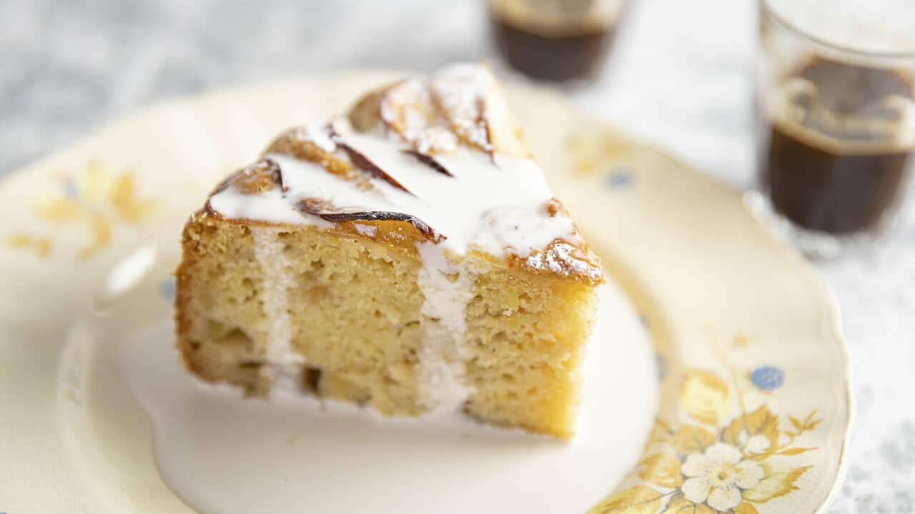 A wedge of pale yellow cake sits on a pretty floral-patterned plate. Cream has been poured over it. Two glasses of coffee can be seen in the background.