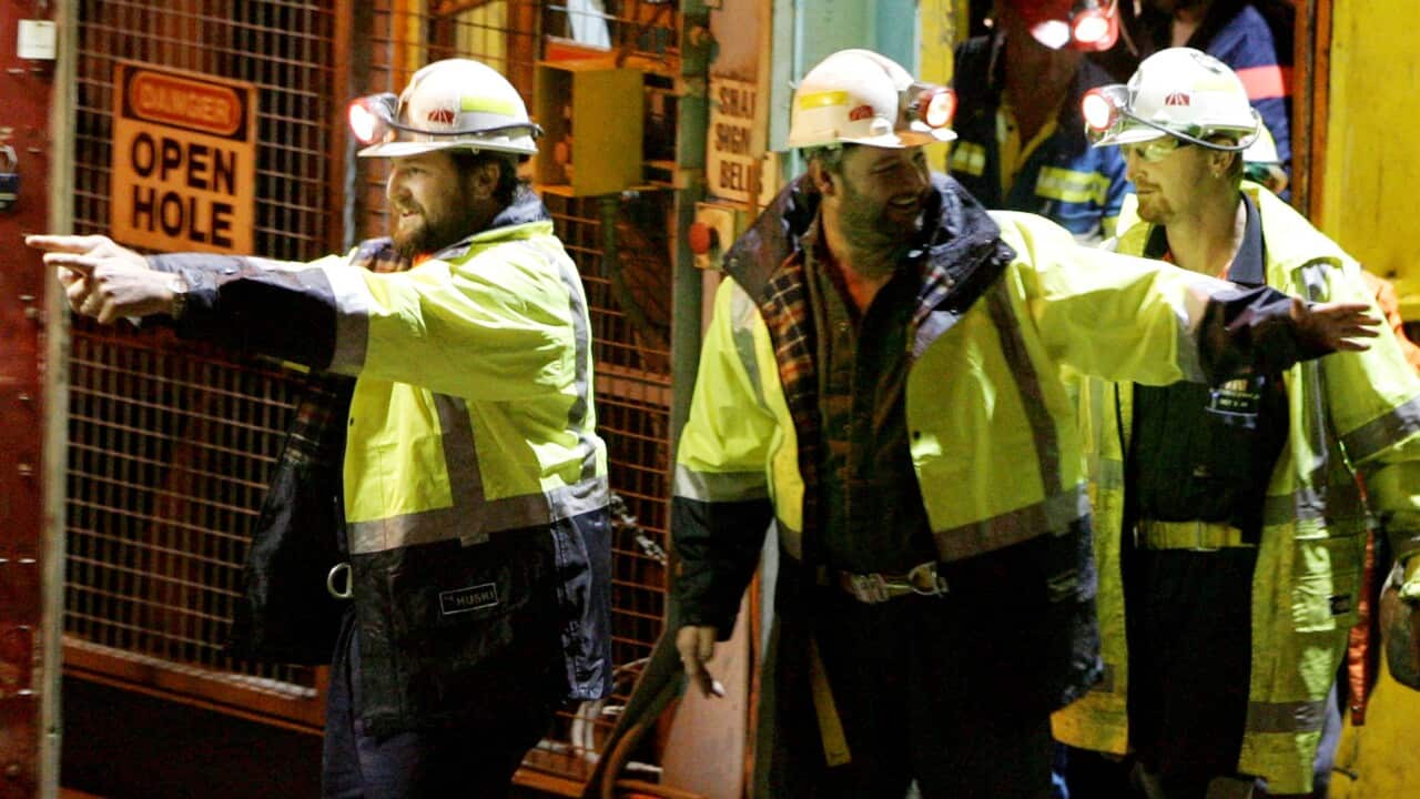 Mining workers wearing hard hats with headlamps and high-visibility jackets stand at a brightly lit worksite
