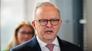 A man in wearing a suit, a tie and glasses speaks with a woman seen in a blurry background