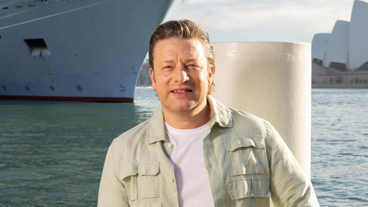 Jamie Oliver wearing a white shirt and green overshirt standing in front of water, a large cruise ship and the Sydney Opera House.