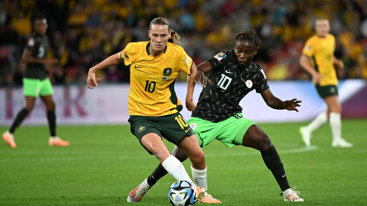 Woman in yellow and green Matildas uniform chases after soccer ball as woman in green and black uniform trails closely behind her on green pitch.