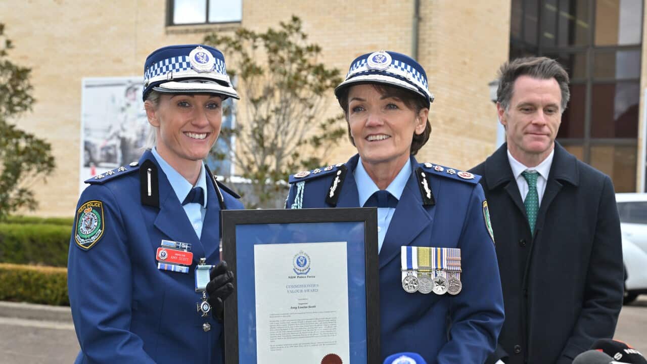A police officer holding a framed award. Another female police officer is next to her, with a man behind them. A row of media microphones is in front.