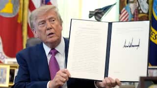 An old man seated at a desk holding up an open document folder displaying a signed official document, with flags and framed items visible in the background.