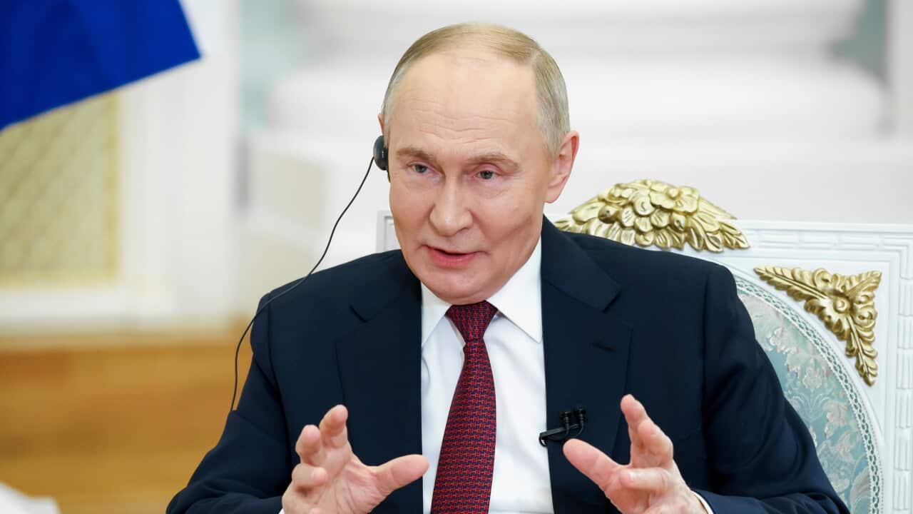 Russian President Vladimir Putin is shown speaking with his hands raised, wearing a black suit and red tie.