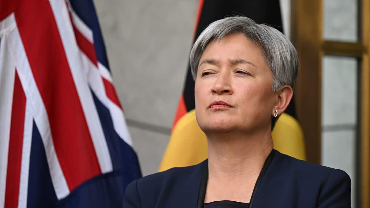 A woman with short, grey hair, wearing a black blazer, stands next to Australian and Aboriginal flags.