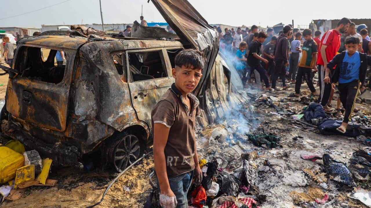 A young boy stands amid ruins. There is a charred car and a crowd of people behind him.