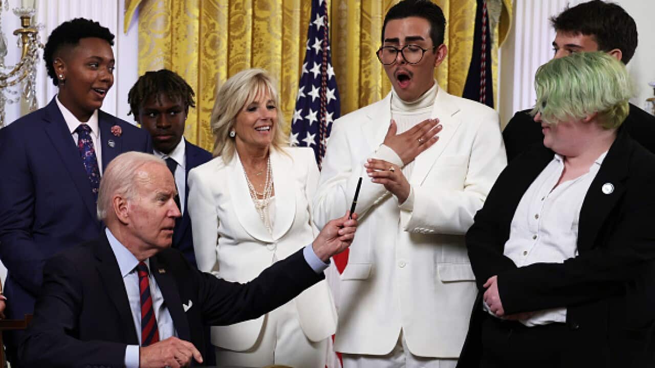 US President Joe Biden sits at a desk in the Oval Office with a group of people standing near him.