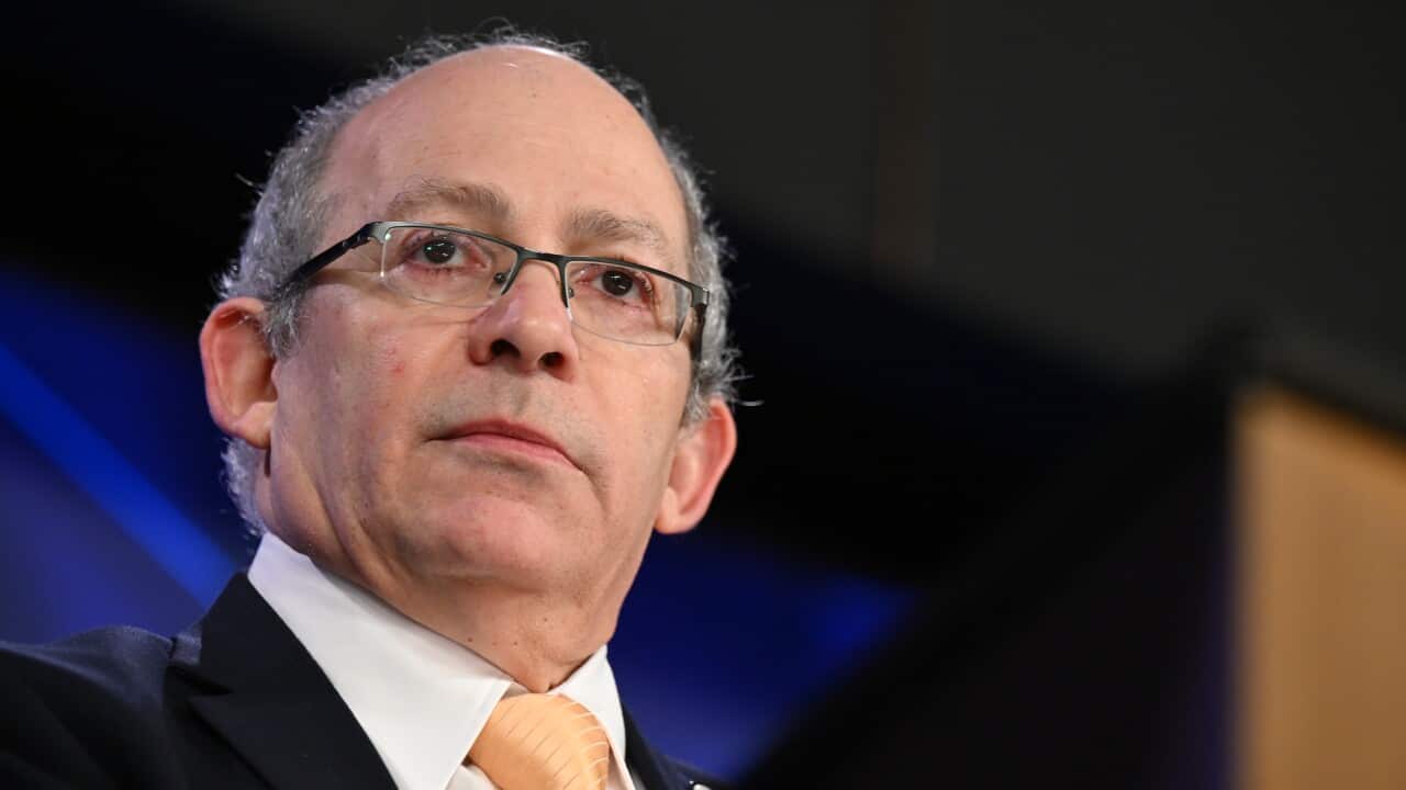 A man wearing a black suit, white shirt and yellow tie. He is wearing glasses and is balding.