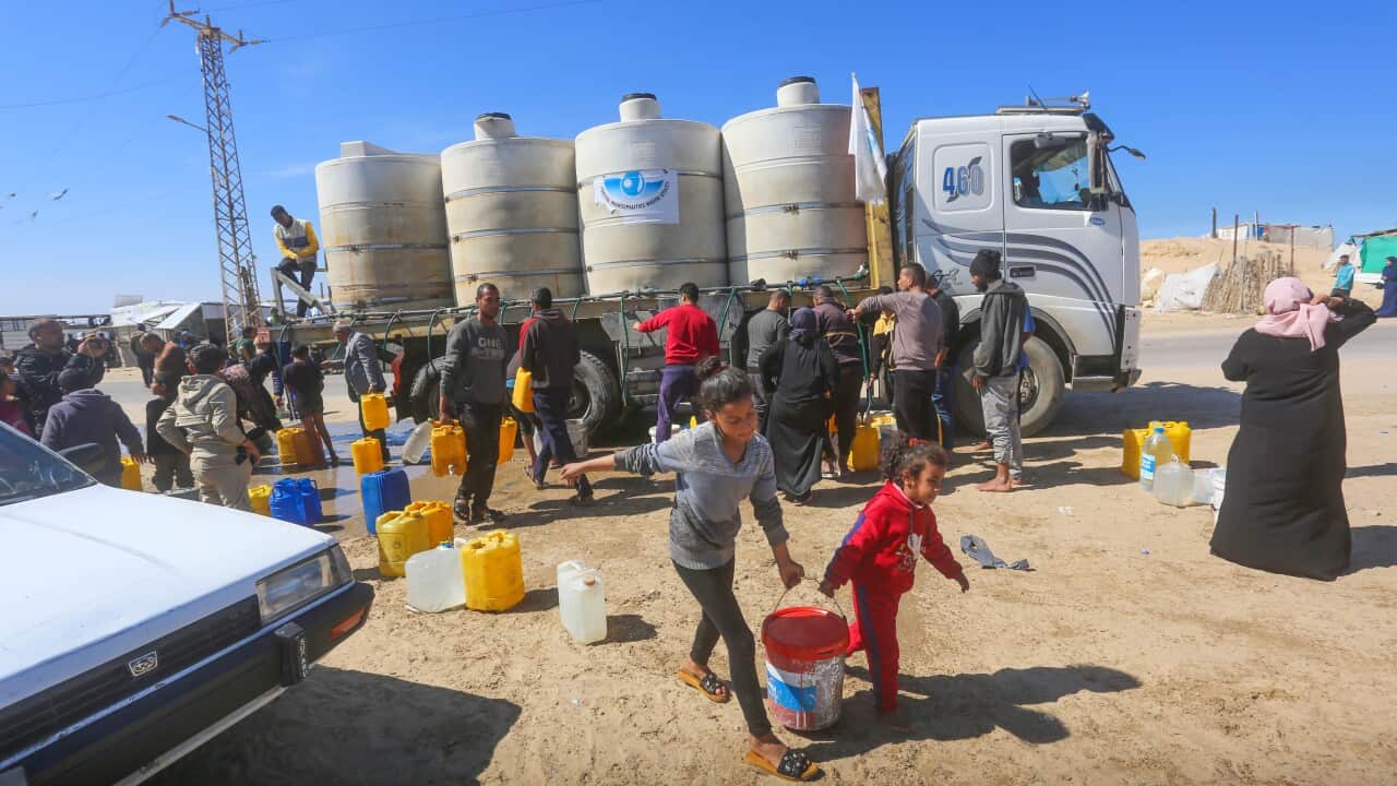 Two children carry a bucket of water with people around a truck delivering aid in the background.