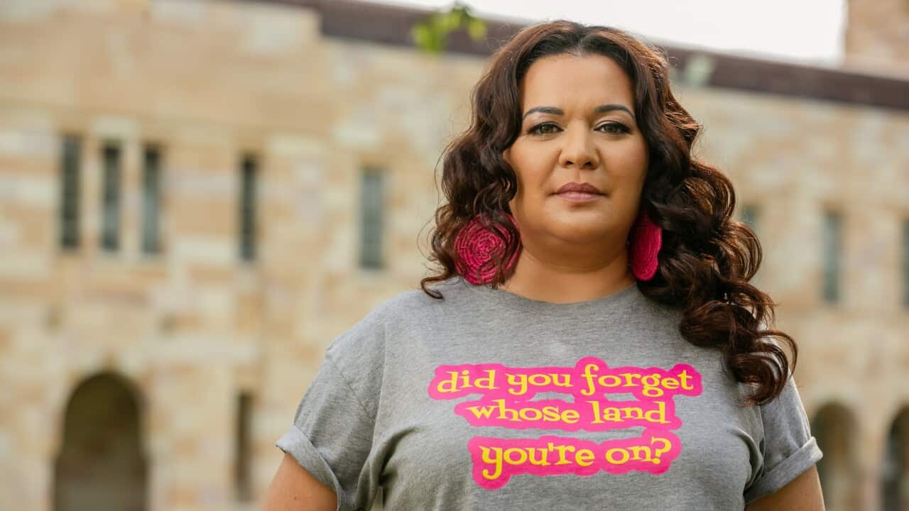 A woman with brown hair standing outdoors and looking at the camera. She is wearing big, pink earrings and a grey t-shirt. The yellow and pink text on her t-shirt reads: did you forget whose land you're on?