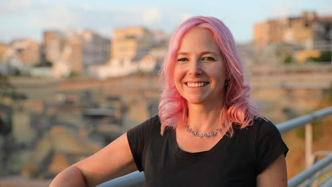 A smiling woman stands beside a rail. A city can be seen behind her.
