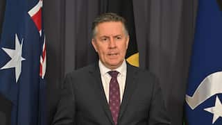 A man in a dark suit and patterned tie speaks at a press conference, standing in front of Australian and Aboriginal flags against a dark curtain backdrop.