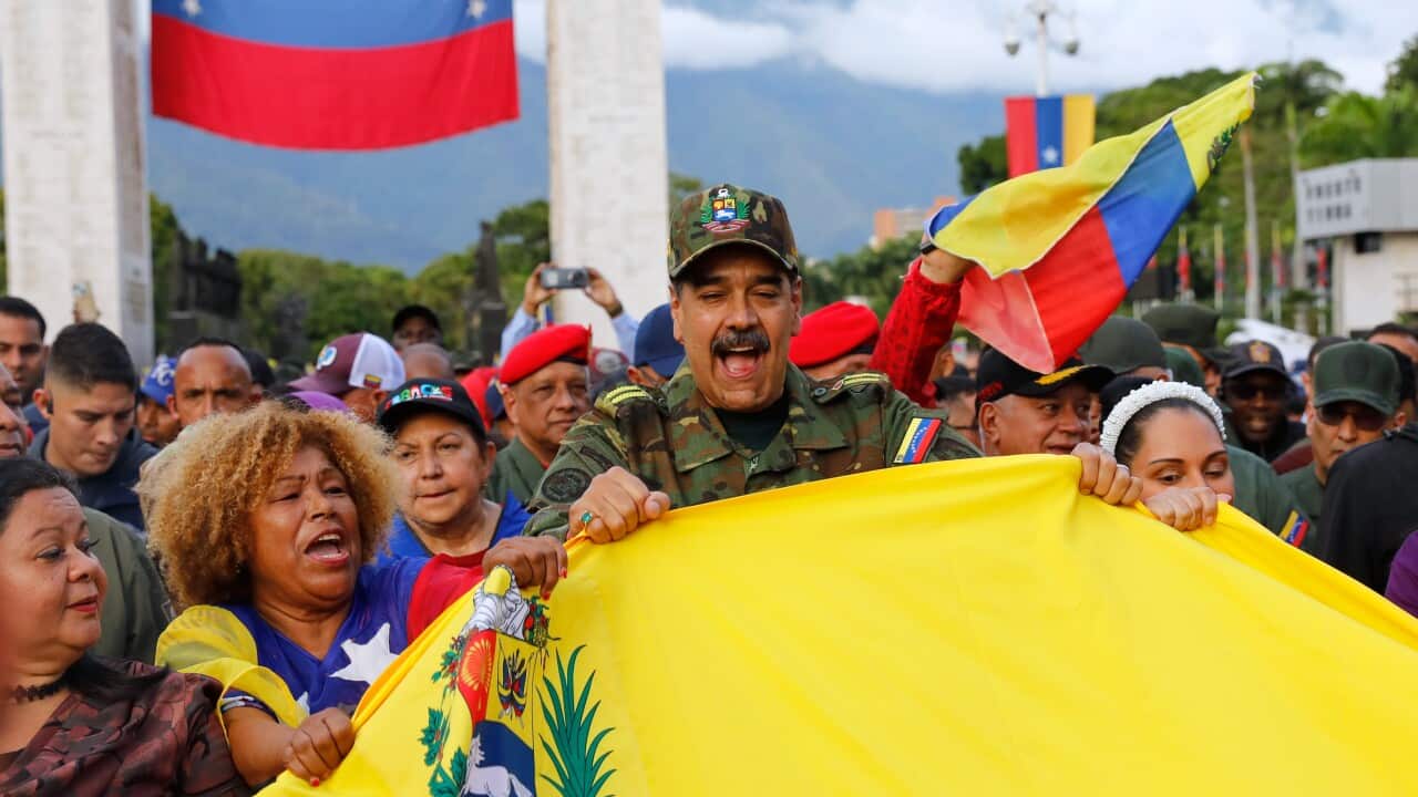 A man in a military uniform holds a yellow cloth with an emblem as he is surrounded by a crowd holding Venezuelan flags in the background.