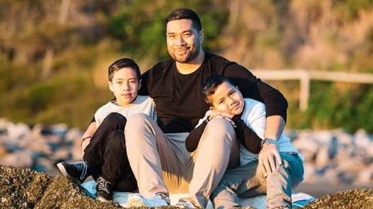 Vae sitting on a hill with his arms around his sons shoulders.