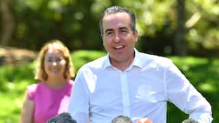 A man wearing a white shirt speaks at a press conference with microphones set up in front of him, and a woman smiling in the background on his right