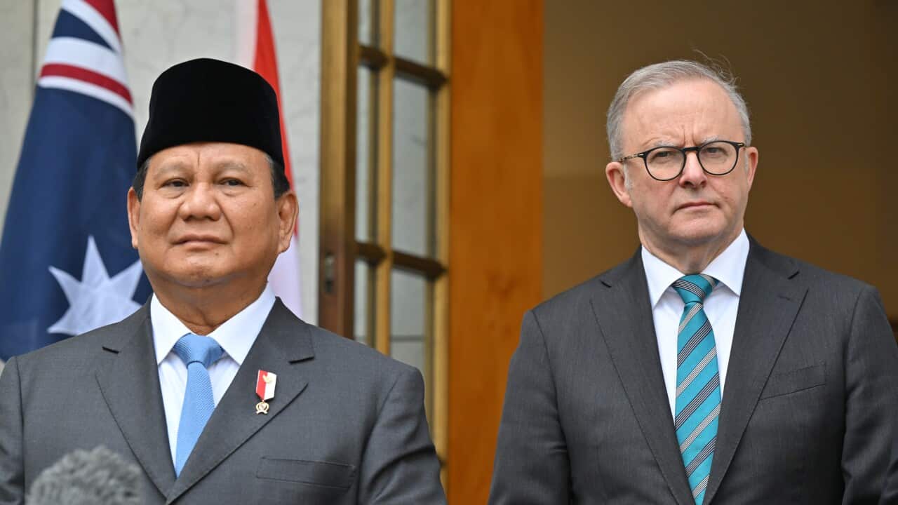 Two men in grey suits stand next to each other. The man on the left is wearing a traditional peci on his head.