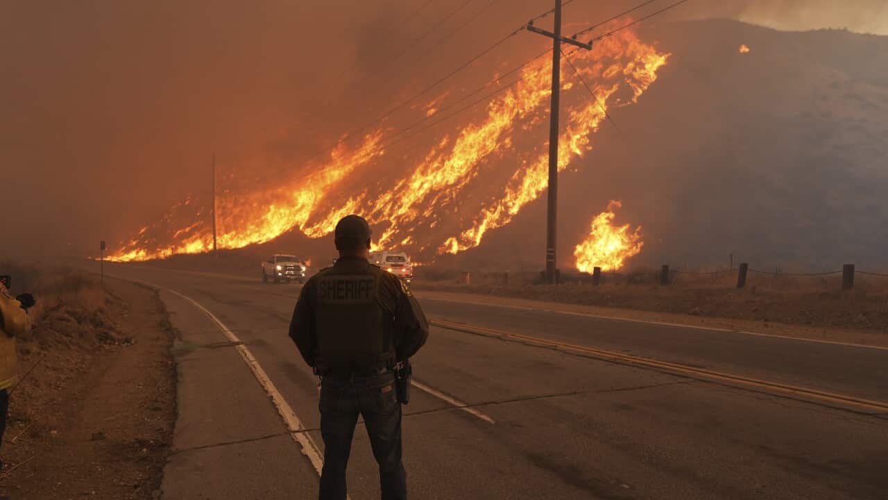 A man stands by the roadside, watching flames burning in the distance.