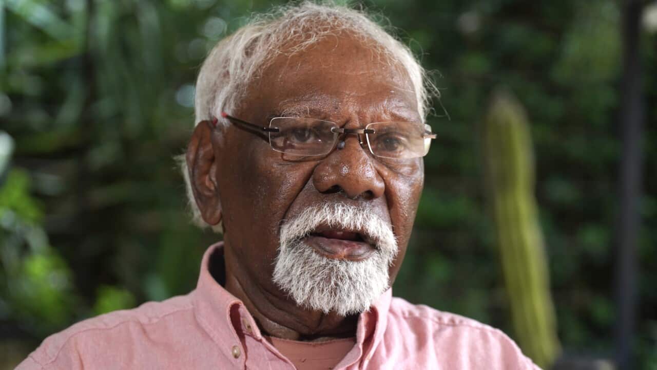 A close-up portrait of an elderly man with short white hair, a white goatee, and glasses, wearing a pink button-down shirt against a blurred green foliage background.