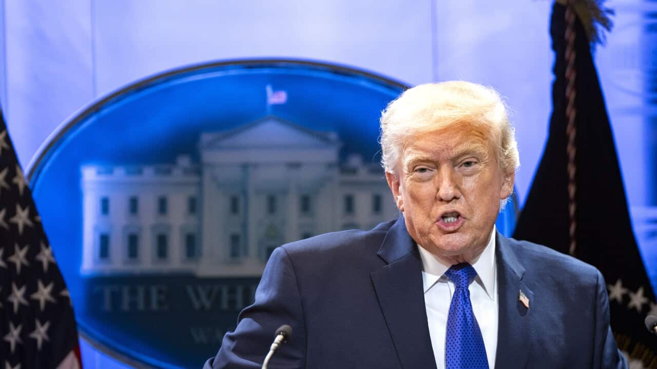Donald Trump, wearing a blue blazer with an American flag pin on it, standing at a podium and microphone, in front of a White House symbol and American flags.