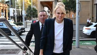 A woman and a man dressed in black blazers and white shirts ascend a small staircase on a city street. The woman has blonde hair and is smiling. The man is middle aged and wears a black tie.