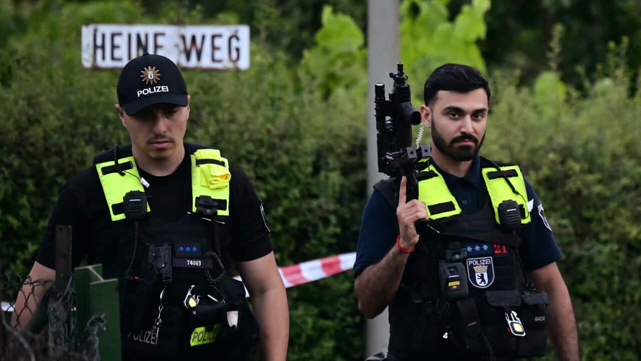 Two police officers wearing bulletproof vests walk near a secured perimeter. The police officer on the left is looking down as he walks, and the other police officer is holding an automatic weapon in his right hand as he walks.