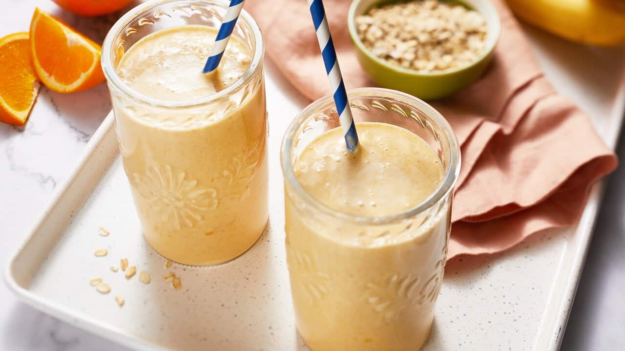 Two glasses with a leaf pattern etched on the side stand on a tray. The glasses hold a golden smoothie mixture. Each has a blue and white striped straw in it. Orange segments and a banana can be seen behind the tray.
