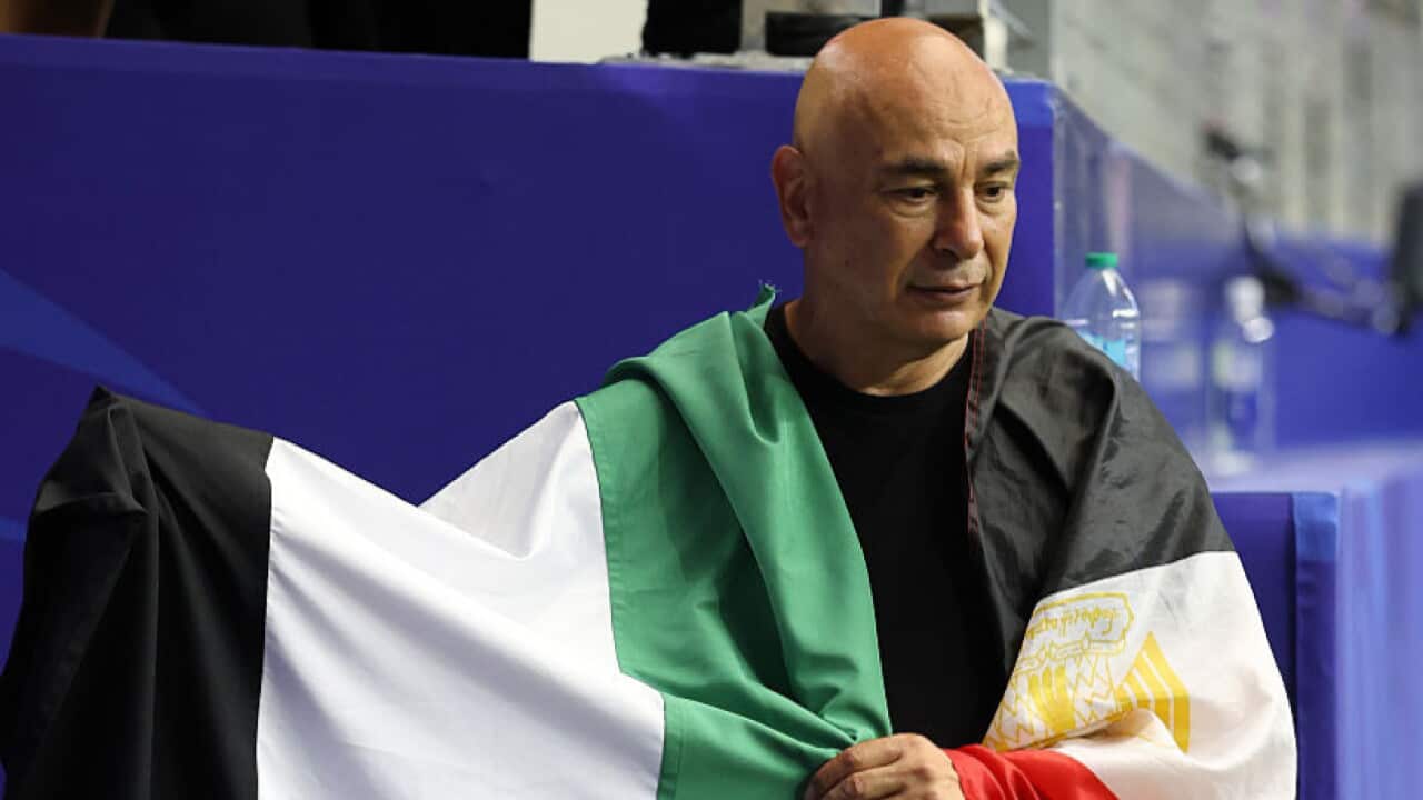 Egypt's coach is draped in Palestine's flag, coloured red, white, green and black, with the blue stadium stands behind him.
