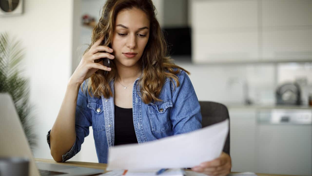 A woman on the phone while seated at a desk, holding a bill in her hand.