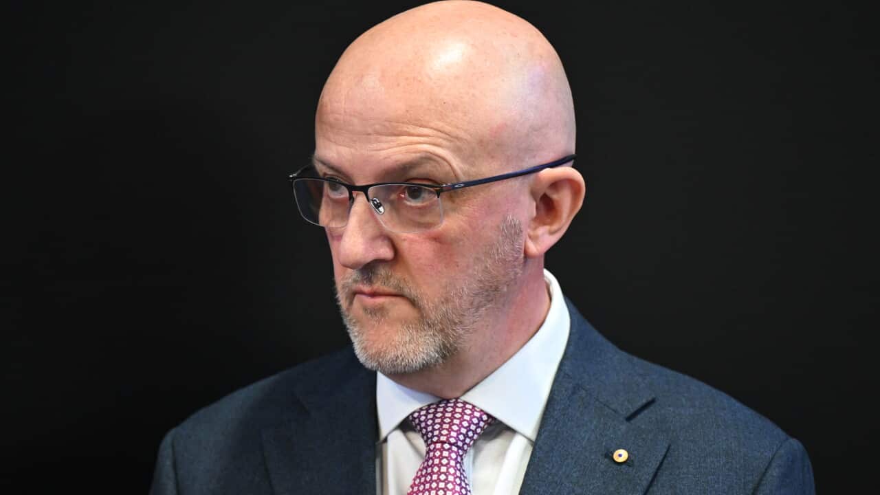 A middle-aged bald man in a blue suit and red patterned tie stands against a black background. He does not look pleased.