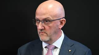 A middle-aged bald man in a blue suit and red patterned tie stands against a black background. He does not look pleased.