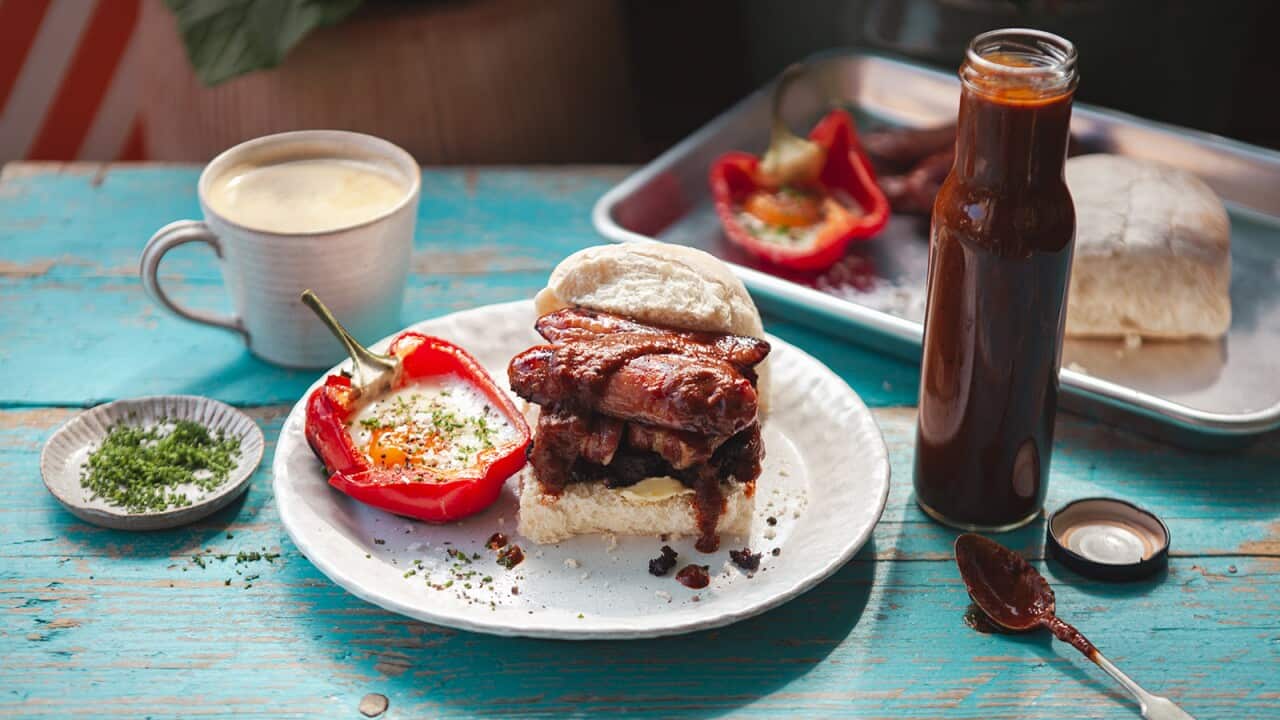 A filled bread roll sits on a white plate, alongside a half capsicum with a cooked egg in it. A cup of coffee sits behind the plate. A tall bottle of brown sauce sits beside the plate. Behind, sitting on the same blue wooden tabletop, is a tray with another bread roll, with fillings and a capsicum half sitting beside it.