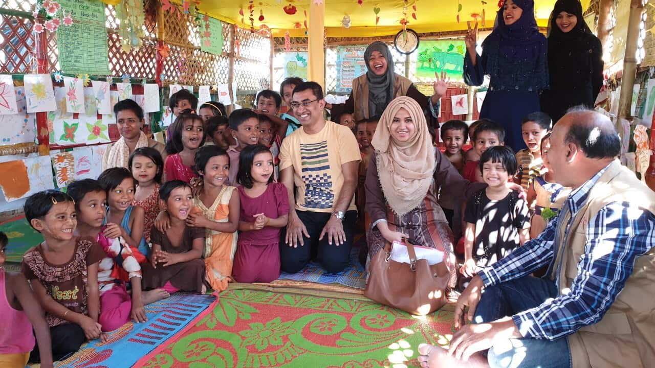 A group of children sit on the floor with some adults smiling on a colourful carpet.