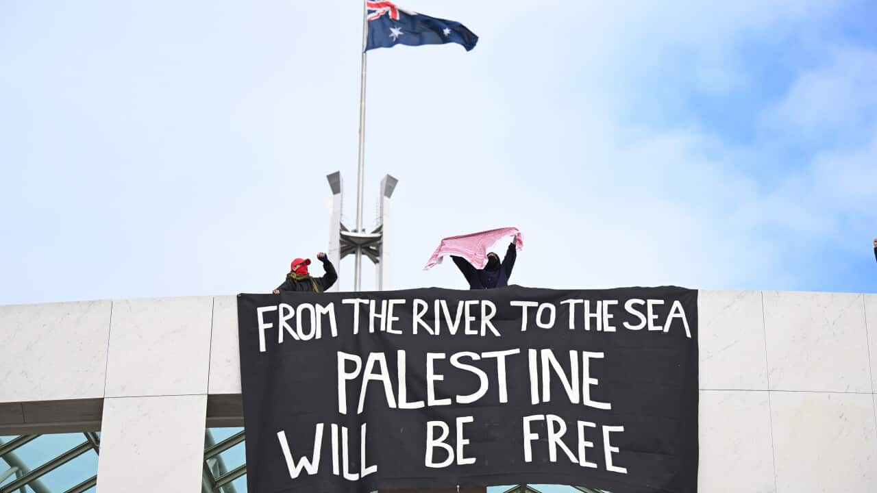Pro-Palestinian activists stand above a large black banner with white text reading "FROM THE RIVER TO THE SEA, PALESTINE WILL BE FREE", which is draped over the edge of a portico.