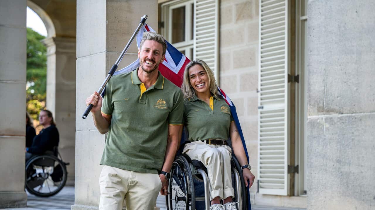 Man holding Australian flag stands next to a woman in a wheelchair in front of a sandstone building