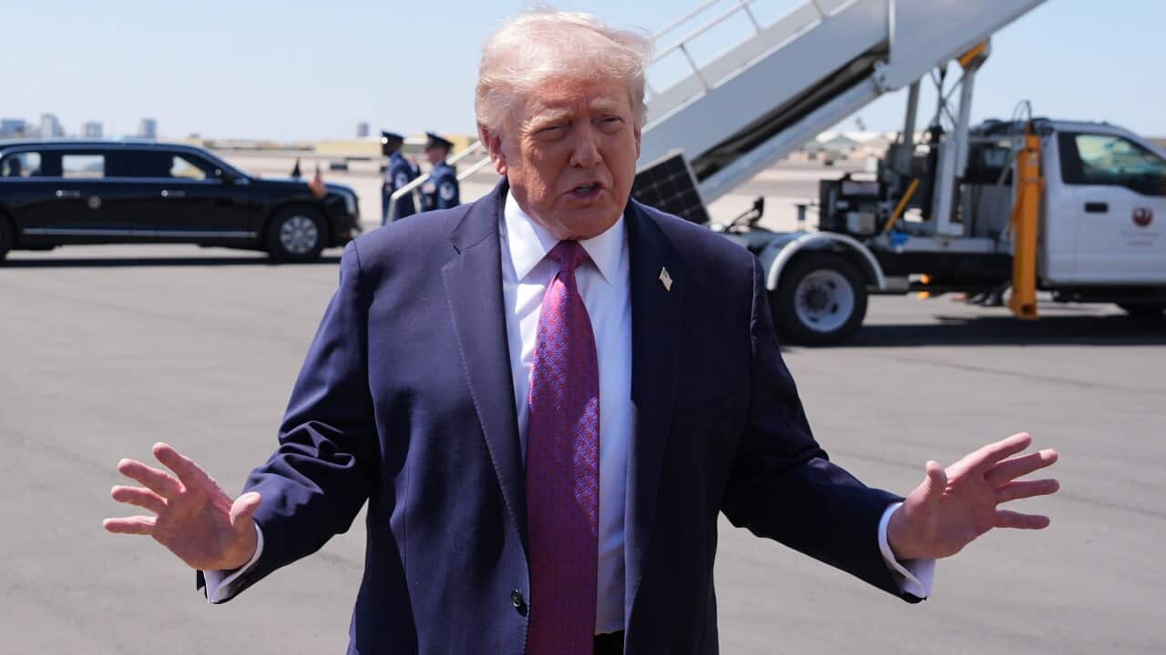 US president Donald Trump stands at an airport. He is dressed in a navy blue suit with a red patterned tie and a US flag pin on his lapel. His hands are outstretched as he talks to reporters. In the background, a motorcade awaits.