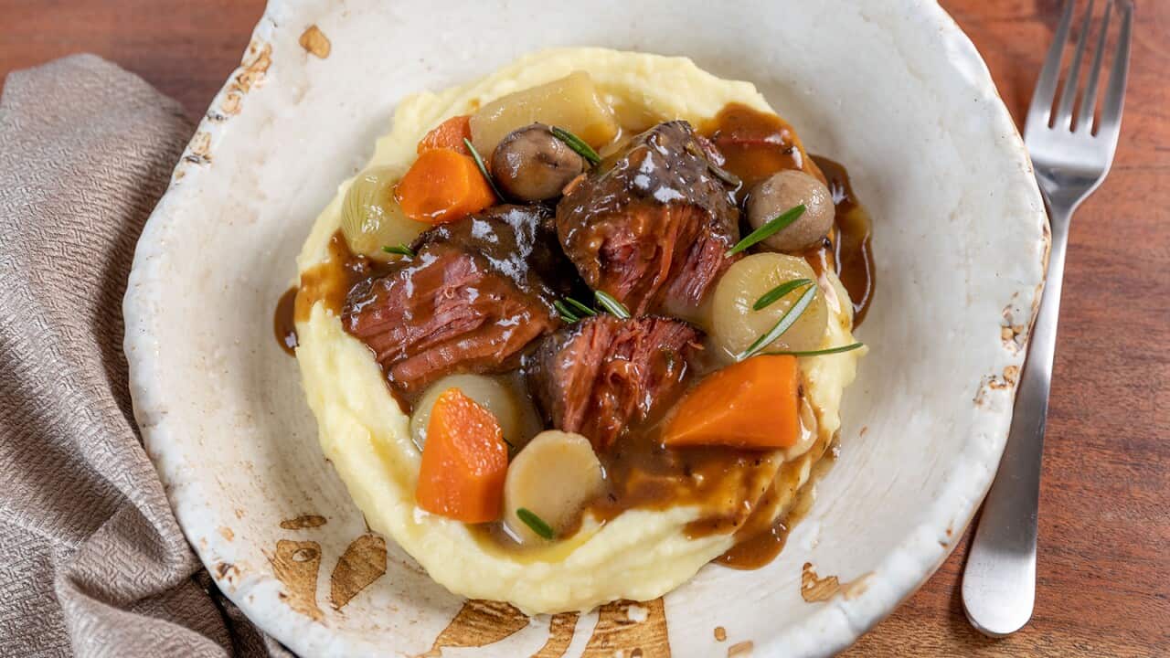 Corn beef pieces, vegetables and bravy sit on a bed of mash in a wide, unevenly shaped white bowl. A fork sits to one side, with a napkin on the other side of the bowl.