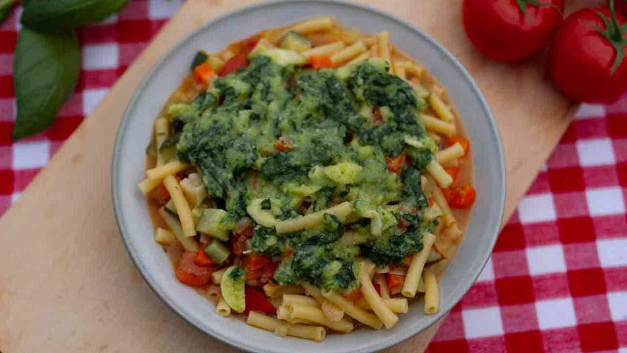 A bowl of soup with tomato and pasta, topped with green sauce, sits on a table.