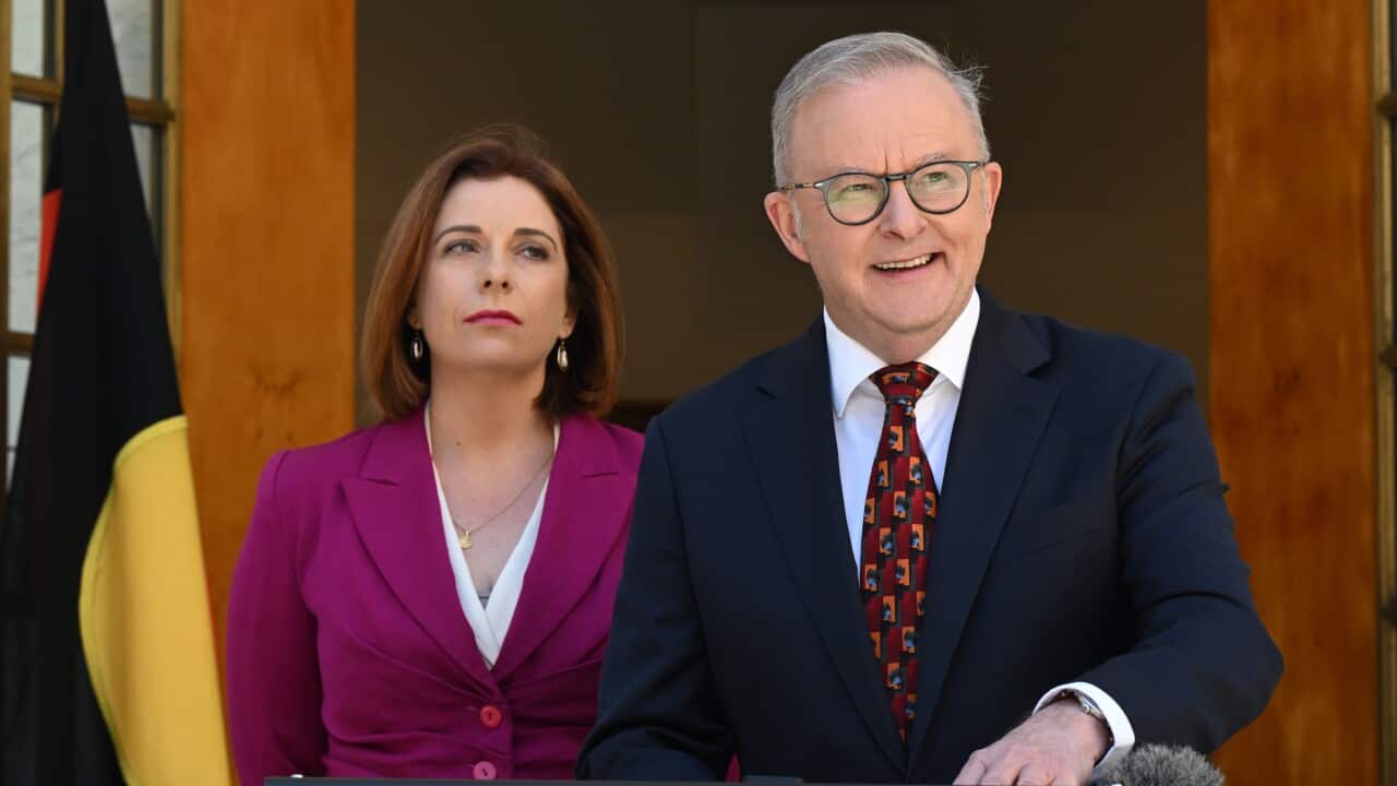 Prime Minister Anthony Albanese speaks at a lectern alongside Communications Minister Michelle Rowland, positioned between the Australian Aboriginal Flag and the Torres Strait Islander Flag.