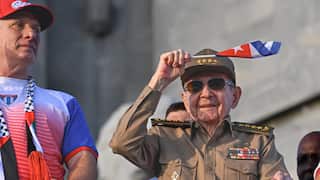 An elderly man in a military uniform smiles and waves a small flag over his head. A man wearing a red and blue t-shirt and a red hat is standing beside him.