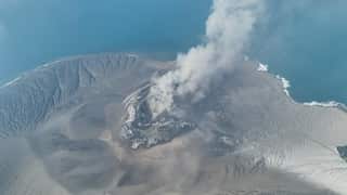 An aerial view of a volcanic island emitting a thick plume of smoke and steam from its central crater surrounded by grey, ash-covered terrain and ocean water.